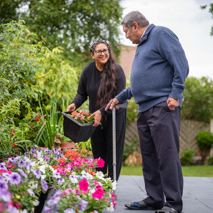 a man with a walking stick is standing next to and pointing towards a flower bed. a woman next to him is smiling and holding some flowers ready to plant where he wants them.